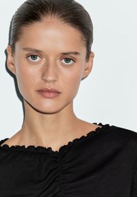 Top nero con scollatura arricciata e dettagli arricciati. Tessuto morbido, indossato da una modella con capelli raccolti, sfondo neutro.