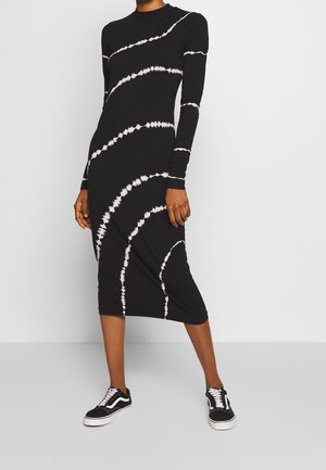 Zwarte jurk met lange mouwen en een wit tie-dye spiraalpatroon, gemaakt van zacht materiaal, knie-lengte, gecombineerd met zwarte sneakers.