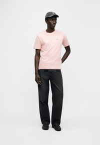 Ροζ βαμβακερό t-shirt με μικρό κεντητό λογότυπο, σετ με σκούρο γκρι παντελόνι φαρδιάς γραμμής και γκρι καπέλο. Απλό φόντο.