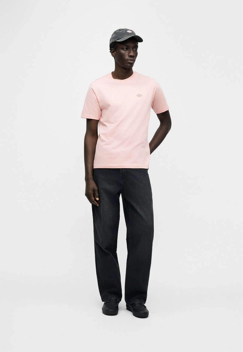 Lichtroze katoenen T-shirt met een klein geborduurd logo, gecombineerd met donkergrijze wijde jeans en een grijze pet. Effen achtergrond.