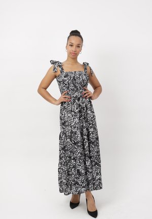 Femme debout, les mains sur les hanches, portant une robe longue sans manches à motifs floraux noir et blanc et des talons pointus noirs sur fond blanc.