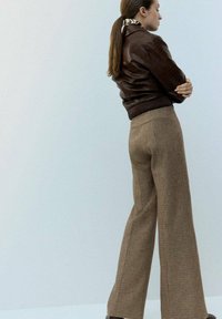 Mujer con chaqueta de cuero marrón y pantalones de pierna ancha de cuadros marrones, de pie de lado con los brazos cruzados contra un fondo claro.