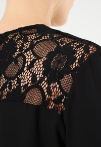 Svart blus med en genomskinlig spetspanel på baksidan med blommönster och olika cirkulära former. Slät tyg på huvudpartiet.