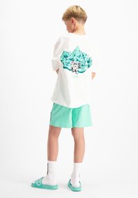 Witte t-shirt met korte mouwen met een grafisch ontwerp in teal op de achterkant. Gecombineerd met mintgroene shorts, witte sokken en aqua slippers.