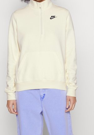 Personne portant un sweat-shirt Nike crème à demi-fermeture éclair avec une poche avant et un pantalon violet clair, debout devant un fond uni.