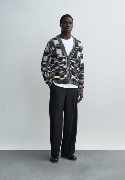 Άνδρας που στέκεται φορώντας μαύρο παντελόνι με φαρδιά μπατζάκια, μαύρη και λευκή ζακέτα με σχέδιο πάνω σε λευκό μπλουζάκι, μαύρα παπούτσια, μπροστά σε απλό φωτεινό φόντο.