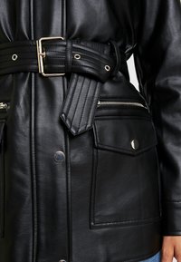 Chaqueta de cuero negro con cintura con cinturón, bolsillos con cremallera y botones a presión. Presenta una textura suave y detalles metálicos.