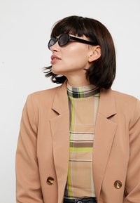 Blazer marrone chiaro con doppio bottone sopra una camicia a quadri multicolore nei toni del beige, verde e marrone. Occhiali da sole rotondi neri completano il look.