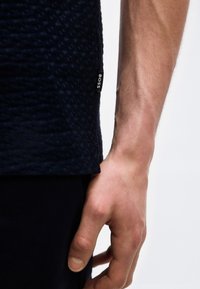 Κοντινή λήψη του βραχίονα και του χεριού ενός ατόμου δίπλα σε ένα σκουρομπλε πουκάμισο με υφή, με ορατή ετικέτα της μάρκας "BOSS" στη ραφή του πλευρού.