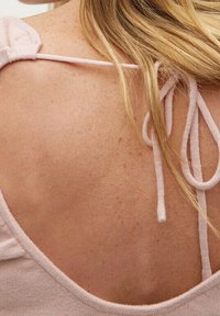 Blondes Haar liegt auf dem sommergefleckten oberen Rücken einer Frau, die ein blassrosa Oberteil mit dünnen Trägern trägt, die an der Schulter gebunden sind.