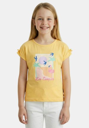 Fille souriante aux longs cheveux blonds portant un t-shirt jaune avec des palmiers, des oiseaux, un flamant rose et le texte "Lee Cooper", assorti d'un pantalon blanc.