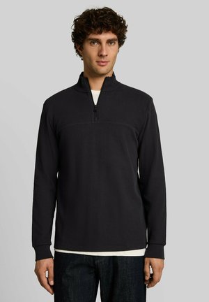 Mann mit lockigem dunklem Haar trägt einen schwarzen Langarm-Halbzip-Pullover über einem weißen Hemd und steht vor einem einfarbigen grauen Hintergrund.