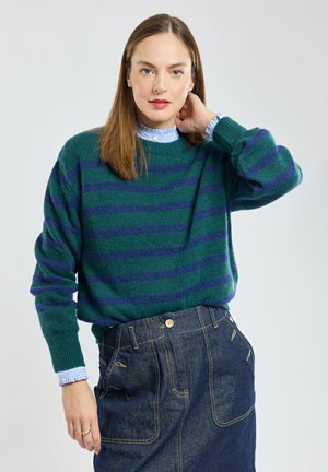 Femme aux cheveux bruns et raides portant un pull rayé vert et marine par-dessus une chemise bleu clair à volants et une jupe en jean foncé taille haute.