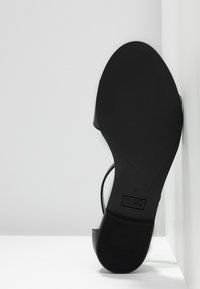 Semelle noire d'une sandale femme à talon bloc appuyée contre un mur blanc, montrant la taille 37 et le logo de la marque Hugo Boss.
