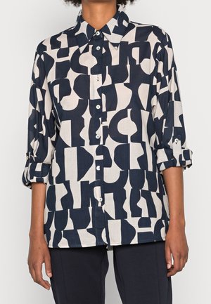 Vrouw draagt een blouse met lange mouwen en knopen, met een zwart-wit abstract geometrisch patroon, mouwen opgerold tot aan de ellebogen.