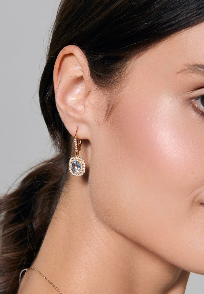 Złota kolczyk w kształcie obręczy z niebieskim kamieniem otoczonym halo z przezroczystych kamieni, o fakturowanej powierzchni i polerowanym wykończeniu.