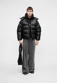 Czarna kurtka puchowa z błyszczącym wykończeniem, skrócony krój i zamek błyskawiczny. W połączeniu z ciemnoszarymi spodniami o szerokich nogawkach oraz czarną torbą na ramię.