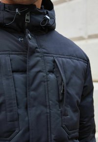 Schwarze isolierte Jacke mit hohem Kragen, verstellbarer Kordelzugkapuze und Reißverschlusstasche auf der Brust. Mit gesteppten Details und Vorderleiste.