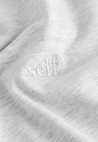Jasnoszara tkanina o miękkiej teksturze z wyhaftowanym logo "self." w kontrastującym kolorze nici. Gładki, lekko melanżowy wygląd.