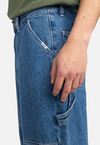Modré džínsy z denimu s bočnou kapsou a malou etikettou. Tkanina má klasickú textúru s viditeľným prešívaním a uvoľnený strih.