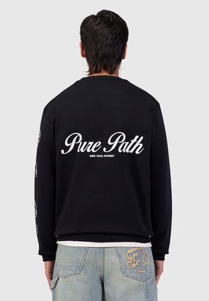 Persoon van achter gezien draagt een zwart sweatshirt met de tekst "Pure Path Own Your Journey" en lichtblauwe spijkerbroek met gouden "PP" borduursel op de achterzak.