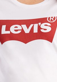Hvit bomulls t-skjorte med en stor rød Levi's-logo med en karakteristisk buet understrek og registrert varemerkesymbol.