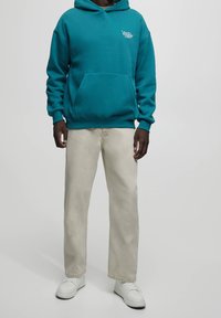 Uomo che indossa un felpa teal, pantaloni beige a gamba dritta e sneakers bianche, in piedi di fronte a uno sfondo grigio chiaro uniforme.