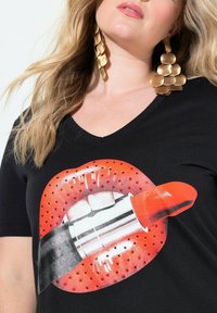Czarny t-shirt z grafiką czerwonych ust i szminki, o błyszczącym wykończeniu, ozdobiony czerwonymi kropkami wokół ust.