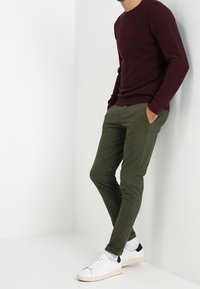 Homme portant un pull bordeaux, un pantalon slim vert olive, des chaussettes blanches et des baskets blanches avec des accents noirs au talon, appuyé contre un mur blanc.