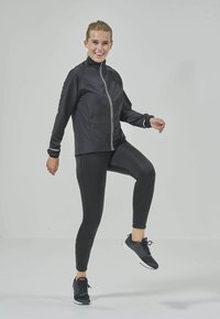 Svart idrottsjacka med vit dragkedja, åtsittande svarta leggings och svarta sneakers. Slät tyg, minimalistisk design och inga synliga mönster.