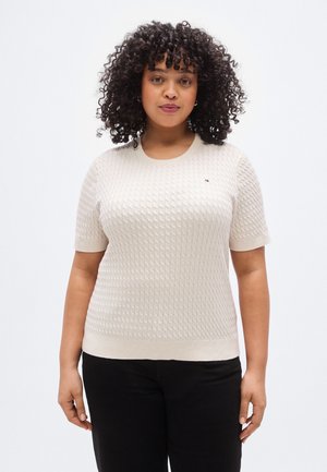 Femme aux cheveux bouclés portant un pull crème texturé à manches courtes et un pantalon noir, faisant face à un fond clair uni.