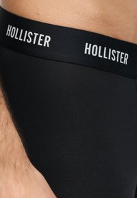 Κοντινή λήψη της ζώνης εσώρουχου μαύρου ανδρικού ρούχου με λευκό κείμενο "HOLLISTER" στο ελαστικό, φορεμένο σε γυμνό δέρμα.