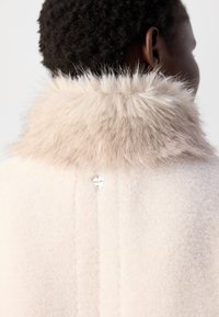 I capelli ricci e corti contrastano con un cappotto di lana beige chiaro con un colletto in pelliccia morbida e un piccolo cartellino metallico sul retro.