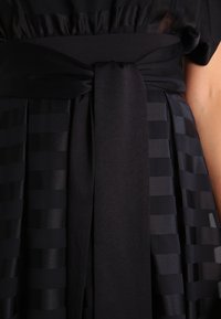 Černé šaty s lesklým saténovým povrchem, zdobené vodorovnými pruhy, s páskem uvázaným v pase a splývavou sukní.