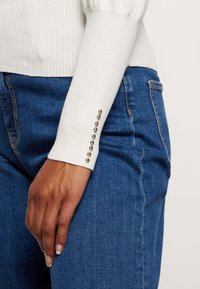 Weißer gerippter Pullover mit dekorativen Nieten am Ärmel, kombiniert mit blauen Denim-Jeans. Nahaufnahme von Ärmel und Handposition.