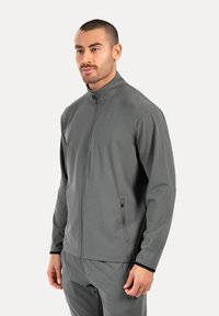 Grå zip-up jakke lavet af blødt materiale, med høj krave, sidelommer med lynlås og en tætsiddende pasform for et atletisk look.