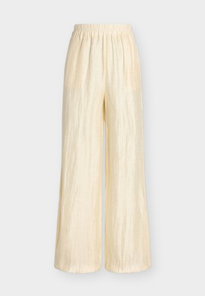 Femme aux cheveux blonds courts portant un pantalon ample de couleur crème et une chemise, un haut blanc, des sandales noires, tenant un petit sac à main noir.