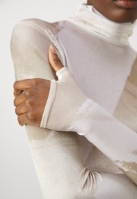 Lange mouwen, hoge hals top van zacht beige stof met een subtiel, textuurpatroon; voorzien van duimgatdetails bij de manchetten.