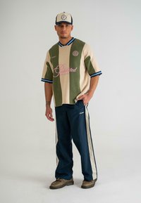 Tricou din jerseu tricotat în culori blocate, bej și măslin, cu decolteu în V, dantelă albastră și logo. Asortat cu pantaloni bleumarin cu dungi albe pe lateral.