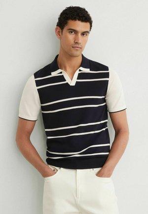 Uomo con capelli ricci che indossa una polo a maniche corte a righe bianche e nere e pantaloni chiari, in piedi con le mani in tasca.