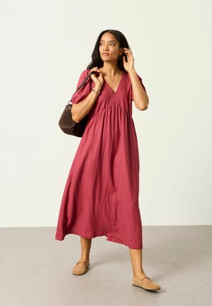 Frau, die ein lockeres, rotes Midikleid und beige flache Schuhe trägt, eine braune Umhängetasche hält und vor einem schlichten hellen Hintergrund steht.