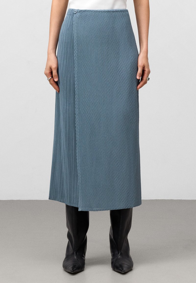 Fälltad, skifferblå midi kjol med sidoknäppning och draperad design, tillsammans med spetsiga svarta stövlar.