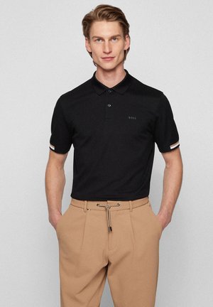 Crna polo majica s ovratnikom, dva gumba i BOSS logotipom. Ima prugaste detalje na rukavima. Uparena s svijetlosmešavim hlačama.