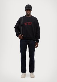Felpa nera con testo ricamato rosso "Ogni sorriso che fingi", abbinata a jeans blu scuro e sneakers beige. Caratterizzata da una tracolla e un berretto nero.
