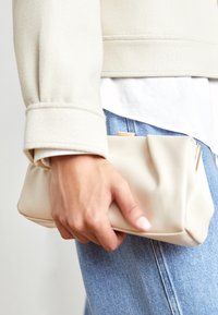 Beige kabelka vyrobená z měkkého materiálu, s nařaseným designem, držená v ruce, s viditelnými modrými džínami v pozadí.