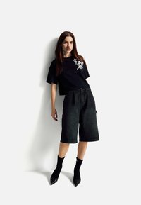 Černé zkrácené tričko s bílým grafickým motivem, kombinované s volnými černými džínovými šortkami, černými ponožkami a černými lodičkami s hrotem.