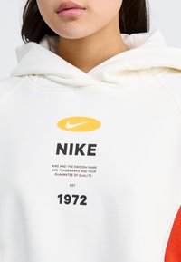 Hoodie blanc avec un logo Nike jaune et du texte noir. Présente un design confortable, une capuche à cordon et des accents rouges sur les manches.