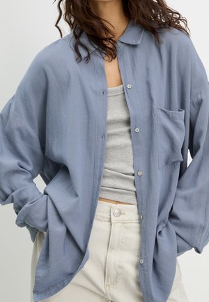 Woman wearing oversized light blue buttoned shirt over gray ribbed tank top and off-white pants, posing with hands in pockets.