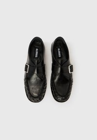 Mustast nahast paari loafereid hõbedaste pandlatega ja ALOHAS kaubamärgiga siltidega sees, esitatud valgel taustal.