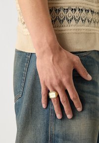 Gylden metallring med et rektangulært, merket topp og teksturerte sider, båret på en hånd, kombinert med en strikket beige topp og denim jeans.